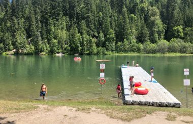 Small green lake with a dock, people mingling about on the grass beach.