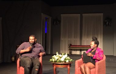 A man and woman sit on chairs on a stage holding microphones.