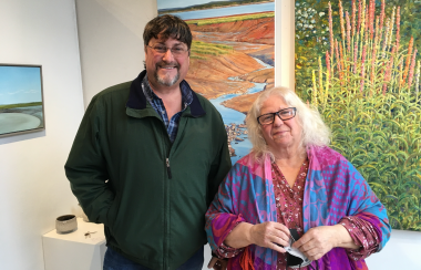 A man and woman standing and smiling, in front of gallery wall full of paintings.