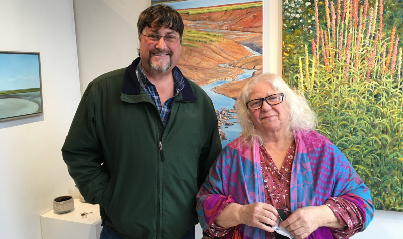 A man and woman standing and smiling, in front of gallery wall full of paintings.