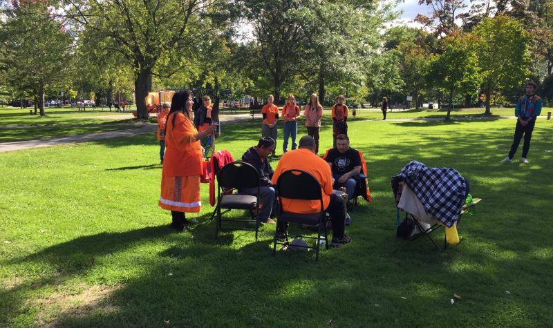 A lady wearing an orange shirt stands while men sit on chairs in a circle
