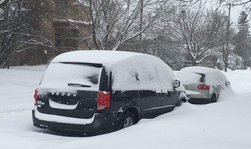 Two vehicles, a navy caravan and a gray sedan, are seen parked at the side of a residential street, partially obscured by heavy snowfall.