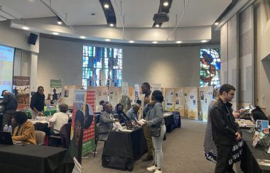 Des dizaines de gens assis et debout autour de kiosques au Salon du livre afro-canadien. Ils sont dans un espace vaste et bien éclairé, on aperçoit des vitraux au fond de la pièce.