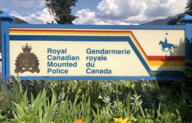 A horizontal sign with the crest of the RCMP in the lower right hand corner. It is outside in a garden of yellow and orange flowers under a cloudy blue sky.