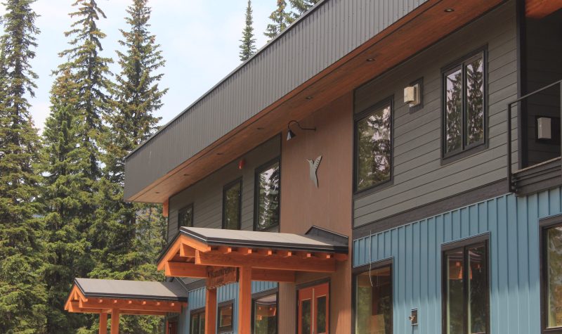 A photo of the lodge. It is a blue and grey building with green trim. Trees in the background.