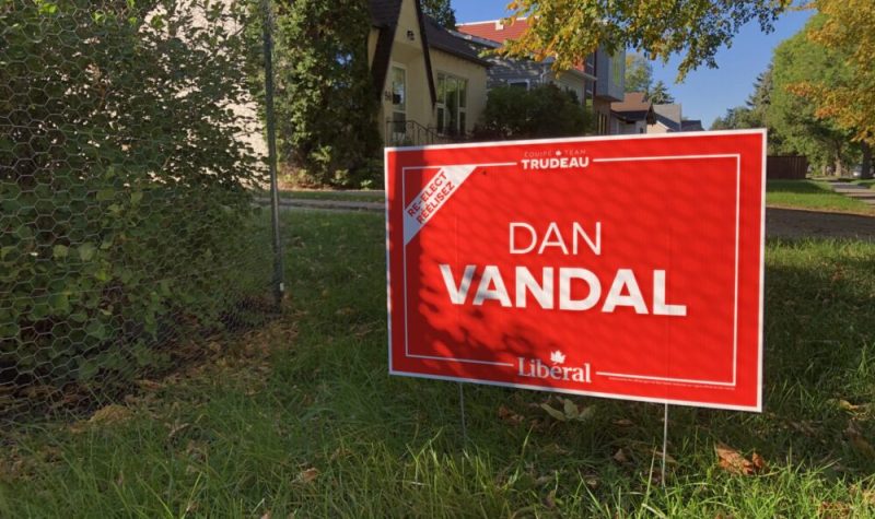 Une affiche rouge de Dan Vandal est plantée dans l'herbe devant une maison dans l'arrière plan.