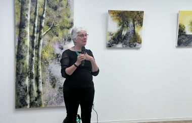 Une femme habillée en noir debout avec un micro et derrière elle des tableaux qui représentent la nature.