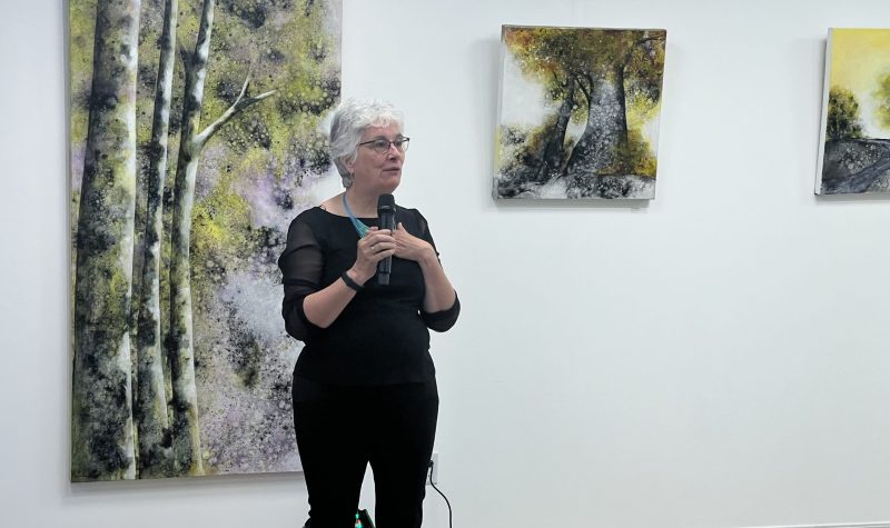 Une femme habillée en noir debout avec un micro et derrière elle des tableaux qui représentent la nature.