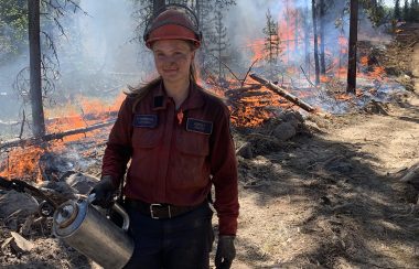 A woman with soot on her face wearing a red shirt with a BC Wildfire Service badges and a nametag reading Ashley Holyk stands in front of a line of fire while holding a canister and wearing an orange hard hat.