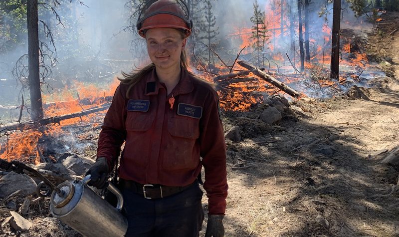 A woman with soot on her face wearing a red shirt with a BC Wildfire Service badges and a nametag reading Ashley Holyk stands in front of a line of fire while holding a canister and wearing an orange hard hat.