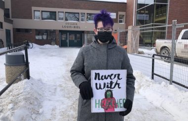Piper est dehors devant l'entrée de son école, iel a les cheveux violets, et tient une pancarte.