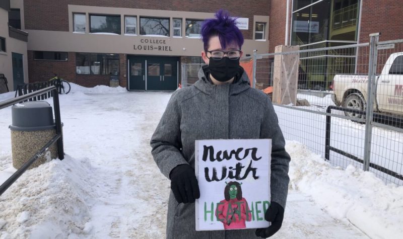 Piper est dehors devant l'entrée de son école, iel a les cheveux violets, et tient une pancarte.