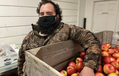 A man wearing a facemask and camouflage jacket is shown leaning on a palette box of red apples.