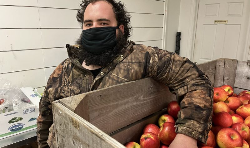 A man wearing a facemask and camouflage jacket is shown leaning on a palette box of red apples.
