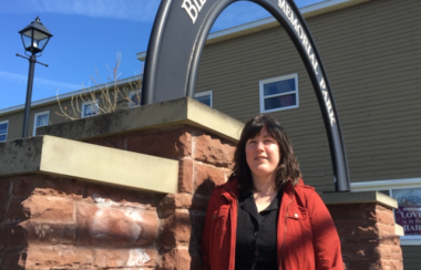 A woman in a red coat standing in front of a gate that reads Bill Johnstone Memorial Park on a sunny day.