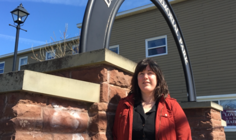 A woman in a red coat standing in front of a gate that reads Bill Johnstone Memorial Park on a sunny day.