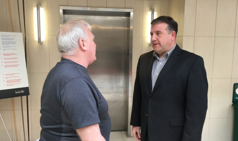 A man in a suit stands facing another man in a grey shirt, who has his back to the camera.