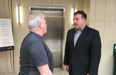 Two men stand face-to-face in what appears to be the lobby of a building. The man on the right wears a dark suit and no tie, and his face is visible. The other man wears a short-sleeved shirt and faces away from the camera. Both of them are light-skinned. The man on the left has white hair and is balding. The man on the right has dark hair.