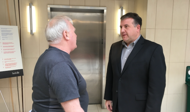 Two men stand face-to-face in what appears to be the lobby of a building. The man on the right wears a dark suit and no tie, and his face is visible. The other man wears a short-sleeved shirt and faces away from the camera. Both of them are light-skinned. The man on the left has white hair and is balding. The man on the right has dark hair.