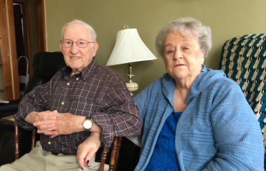 An older man in a plaid shirt sitting beside an older woman in a blue sweater. They are arm in arm, and smiling.