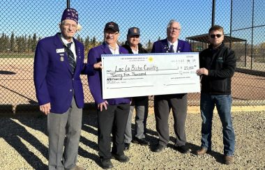 Le maire du comté de Lac La Biche Paul Reutov pose pour une photo avc un chèque géant de 25 000$ en compagnie de quelques représentants du Elks Lodge de Lac La Biche, derrière la clôture du terrain de baseball.