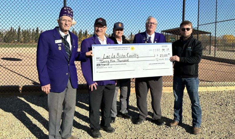 Le maire du comté de Lac La Biche Paul Reutov pose pour une photo avc un chèque géant de 25 000$ en compagnie de quelques représentants du Elks Lodge de Lac La Biche, derrière la clôture du terrain de baseball.