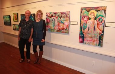 Two people stand in front of a gallery full of colorful art.