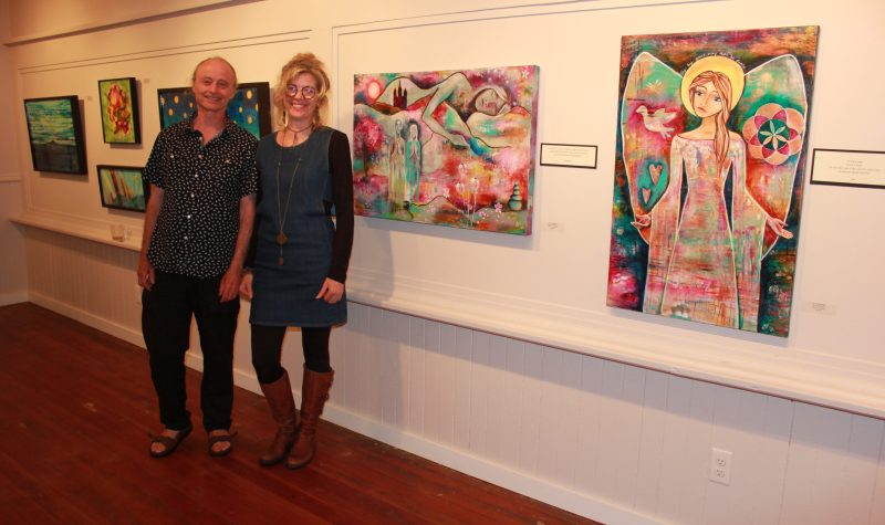 Two people stand in front of a gallery full of colorful art.