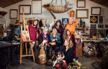 Eight women playfully pose in an art studio surrounded by multi media work.
