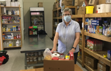 A woman standing in a room surrounded by shelving and a fridge stocked with food. She has her hands on a box sitting on a table in front of her, stocked with dry goods.