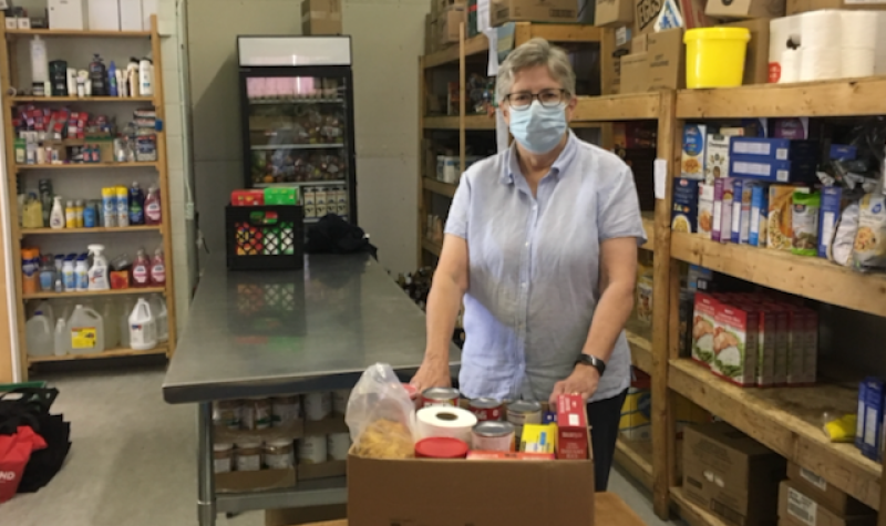 A woman standing in a room surrounded by shelving and a fridge stocked with food. She has her hands on a box sitting on a table in front of her, stocked with dry goods.