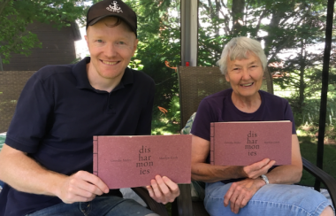 A man in a ballca and a woman with short grey hair sitting in a greenspace holding two books.