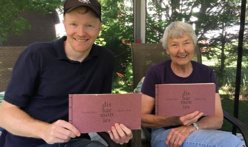 A man in a ballca and a woman with short grey hair sitting in a greenspace holding two books.