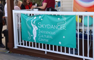 A turquoise banner with the Skydancer Indigenous Cultural Centre and Art Gallery's logo on it, hung on a railing on the front porch of the building. Weather is clear.
