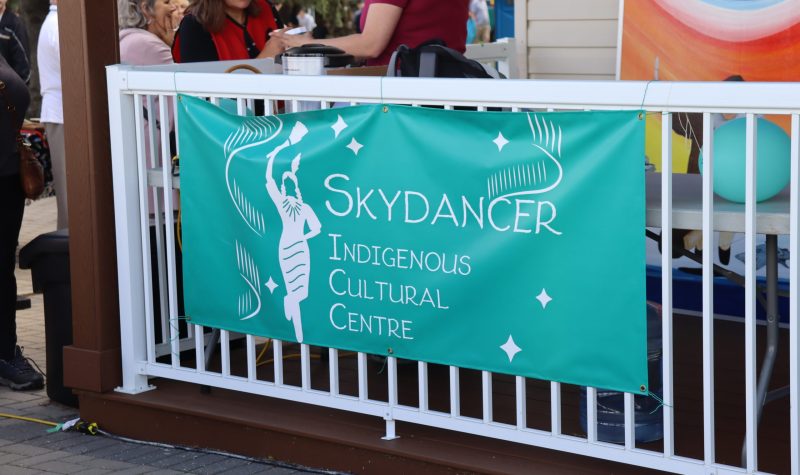 A turquoise banner with the Skydancer Indigenous Cultural Centre and Art Gallery's logo on it, hung on a railing on the front porch of the building. Weather is clear.