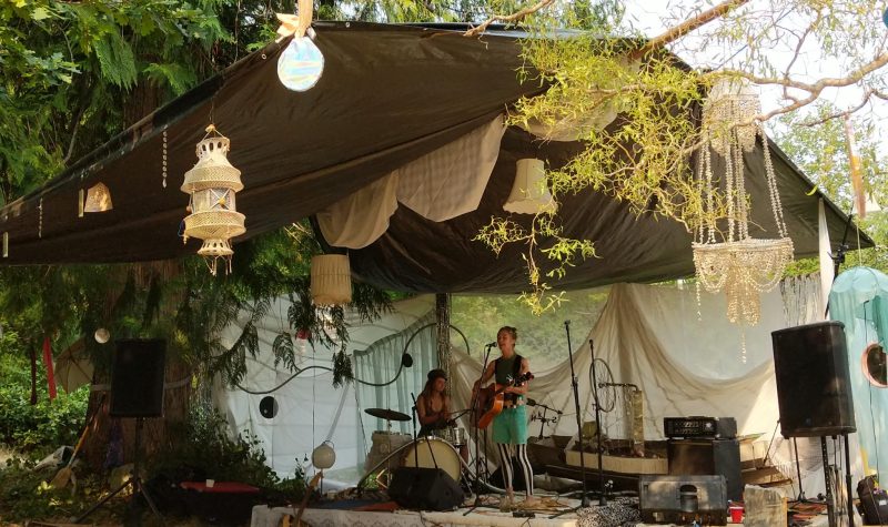 Two musicians perform on an outdoor stage with white adorned chandelier installations surrounding.