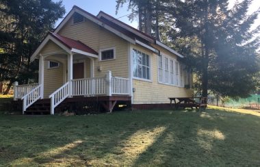 A rural one room schoolhouse painted in soft cream tones with a red asphalt roof and surrounding trees sees the morning sunrise with shadows cast upon a green lawn.