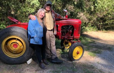 A woman and man stand in front of a 1950's big red tractor with forest backdrop.