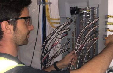 Un homme est en train de mettre en place un panneau électrique, avec une veste réfléchissante et des lunettes de protection.