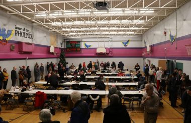 A wide shot of the Bent Arrow Gymnasium with people lined up all the way around the perimeter of the gym waiting for some soup and Bannock.