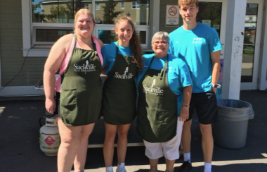 Four people standing in front of a large BBQ on a sunny summer day.