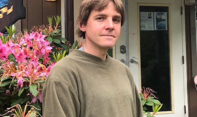 A man in an olive green sweater stands in front of a pink rhododendron bush.