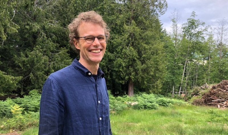 A blonde haired man with glasses wearing a blue shirt smiles in front of a forest background.