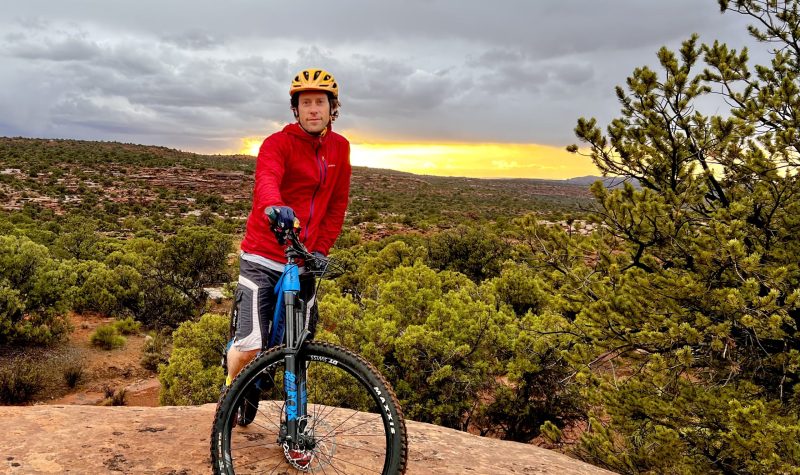 Man in red jacket posing on a mountain bike. Sunset in the background.