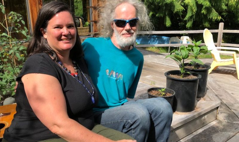 A dark haired woman in a black t-shirt and a man with grey hair and beard wearing sunglasses sit on a deck.