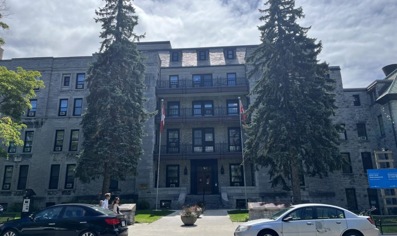 Kingston General Hospital, a tall grey building with two large pine trees framing the entrance. it is a sunny day.