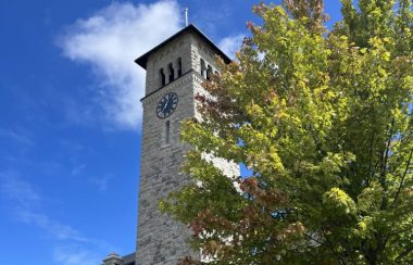 a blue sky with white clouds, looking up at grant hall, a grey stone building with a tall clock tower. a tree partially obscures the view of the building