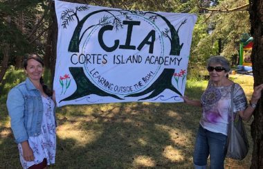 Two women stand on either side of a banner announcing the Cortes Island Academy in a forested setting.
