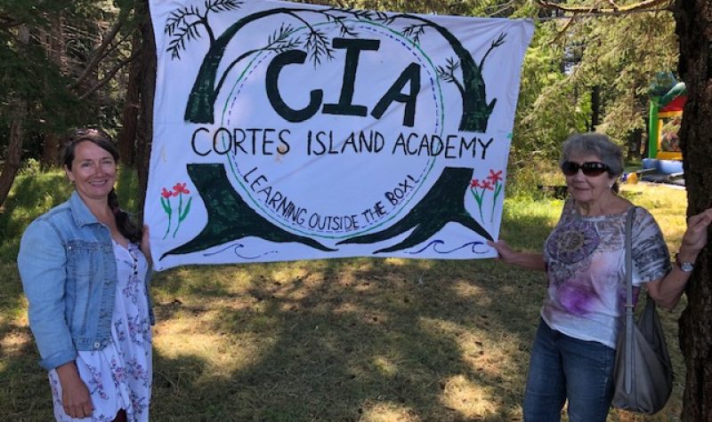 Two women stand on either side of a banner announcing the Cortes Island Academy in a forested setting.
