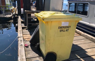A yellow waste bin sits on a dock with boats tied up alongside.